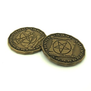 Puede incluir: Dos fichas de metal de color dorado con un diseño de pentagrama. Las fichas tienen una superficie texturizada y un borde elevado.
