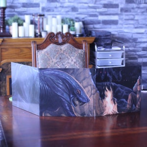 Może przedstawiać: Trójdzielna, składana plansza do gry z ilustracją fantasy. Panel środkowy przedstawia niebiesko-szare stworzenie z ostrymi zębami i pazurami, ogień i postać z mieczem. Panel lewy przedstawia rozmyte zdjęcie ogona stworzenia. Panel prawy przedstawia rozmyte zdjęcie postaci z mieczem.