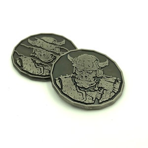 May include: Two silver-colored metal coins with a raised design of a warrior's face wearing a helmet. The warrior has a fierce expression and is wearing armor.
