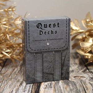 Puede incluir: Una caja gris con textura de cuero y el texto "Quest Decks" en letras negras. La caja también tiene el texto "Emissaries & Infiltrators" en letras negras.