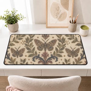 May include: A rectangular desk mat featuring a repeating pattern of brown moths and intricate green and blue acanthus leaf scrolls on a cream-colored textured background. The mat has a thin black border and a flat, smooth surface design.