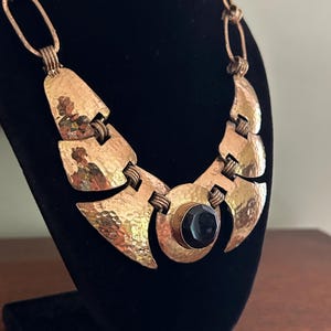May include: A vintage gold-toned necklace with a hammered texture. The necklace features a large, dark, round stone at the center and is displayed on a black velvet bust. The chain is made of oval links.