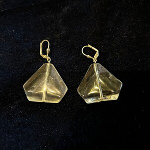 May include: A pair of clear glass earrings with gold-toned metal hooks. The earrings are shaped like a diamond with a faceted design.