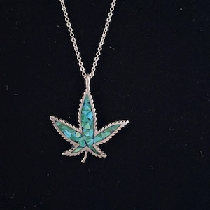 Puede incluir: Un colgante plateado con forma de hoja dentada, incrustado con pequeñas piezas de mosaico azul y verde iridiscente, suspendido de una fina cadena de eslabones de metal plateado a juego.