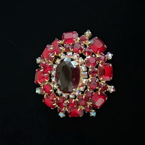 May include: A vintage brooch with a gold-tone setting, featuring a large oval, dark red center stone surrounded by smaller, vibrant red and clear crystals. The brooch has a floral-like design and is set against a black background.