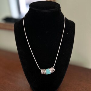 May include: A silver necklace with a turquoise-colored center stone. The necklace is displayed on a black mannequin. The center stone is encased in silver with an ornate design. The necklace is a simple, elegant piece of jewelry.