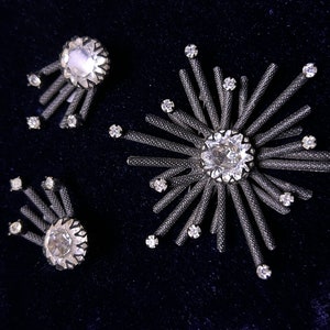 May include: A set of three silver tone brooches with clear rhinestone accents. The brooches are in the shape of a starburst with radiating points.
