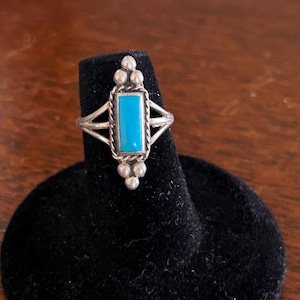 May include: A silver ring with a turquoise stone set in a rectangular shape. The ring has a beaded design on the band.