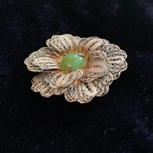 Puede incluir: Broche de flores de filigrana plateada con una piedra central ovalada de color verde claro. El broche tiene pétalos calados detallados y está sobre un fondo oscuro. Una joya de estilo vintage.