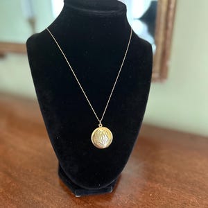 Op de afbeelding: Een gouden medaillonketting met een fijne ketting wordt gepresenteerd op een zwarte fluwelen buste. Het ronde medaillon heeft een gegraveerd ontwerp, mogelijk initialen of een monogram. De ketting wordt gepresenteerd op een donkere houten ondergrond.