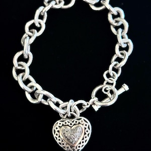 May include: Silver-toned bracelet with a heart-shaped charm. The charm features a gold-toned heart filled with small, sparkling stones, set within a filigree design. The bracelet has a toggle clasp and a chain link design.