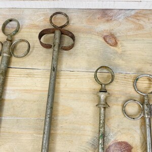 Peut inclure: Quatre outils anciens en m&eacute;tal avec des poign&eacute;es en anneau et de longues tiges, dispos&eacute;s sur une surface en bois. Les outils pr&eacute;sentent des signes d'&acirc;ge, avec un aspect vieilli et terni et une certaine oxydation verte. La conception des outils varie.