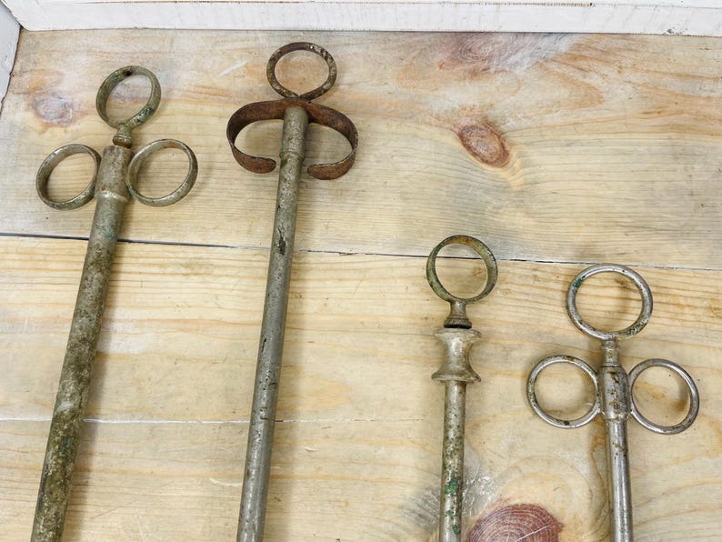 Peut inclure: Quatre outils anciens en m&eacute;tal avec des poign&eacute;es en anneau et de longues tiges, dispos&eacute;s sur une surface en bois. Les outils pr&eacute;sentent des signes d'&acirc;ge, avec un aspect vieilli et terni et une certaine oxydation verte. La conception des outils varie.