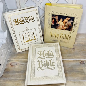 May include: Three vintage Holy Bibles with gold lettering and decorative borders. One Bible is open, displaying text and a small illustration. The other two Bibles are closed, with the title "Holy Bible" prominently displayed on the covers. The Bibles are in various shades of cream and beige.