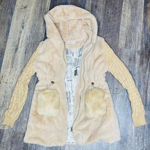 May include: A beige hooded coat with a faux fur body and knitted sleeves. The coat features a drawstring waist, two front pockets, and a zipper closure. The interior lining has a printed design.