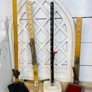 May include: A collection of vintage measuring tools, including wooden rulers, a black skirt marker, and a metal measuring device. The rulers are marked with inch measurements. The tools are displayed against a white, decorative backdrop.