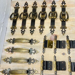 May include: A collection of antique bronze-toned hardware, including door handles and hinges. The handles have a decorative design with a ring pull. The hinges are a mix of bronze and black. The hardware is arranged on a light wood surface.