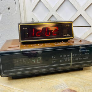 May include: Two vintage digital alarm clocks. The top clock displays "12:02" in red digits, with "Auto Dimmer" text. The bottom clock shows "18:48" and has a radio dial with "Panasonic" branding.