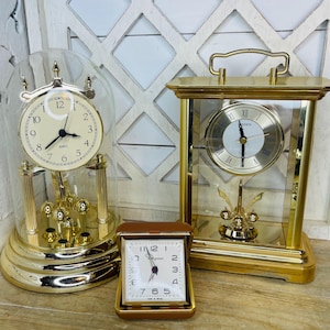 May include: Three vintage clocks in gold tones. One is a quartz clock under a glass dome, another is a rectangular clock with a handle, and the third is a small travel clock in a brown case. The clock faces have Roman and Arabic numerals.