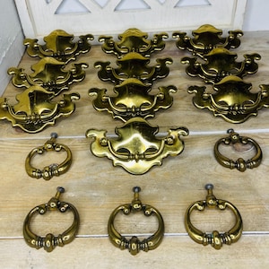 May include: A collection of vintage brass drawer pulls and ring pulls. The drawer pulls have a decorative, curved design, while the ring pulls are circular with ornate detailing. The hardware is arranged on a light-colored wooden surface.