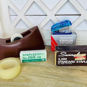 May include: An assortment of office supplies, including a brown tape dispenser with a roll of tape, a blue translucent stapler, and boxes of staples. A roll of clear tape and a box of staples are also visible.