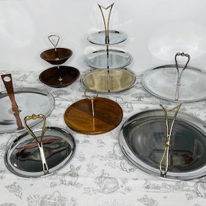 May include: An assortment of serving trays and tiered stands. The trays are oval and round, made of silver, wood, and metal. Some have handles in gold and silver. A three-tiered stand with glass plates and a gold metal frame is also present.