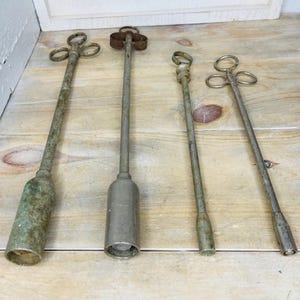 Peut inclure: Quatre outils anciens en m&eacute;tal avec de longs manches et divers motifs sup&eacute;rieurs. Les outils ont un aspect vieilli et patin&eacute;, avec un m&eacute;lange d'argent et de vert. Ils sont dispos&eacute;s sur une surface en bois clair.
