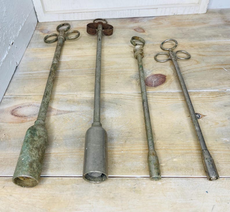 Peut inclure: Quatre outils anciens en m&eacute;tal avec de longs manches et divers motifs sup&eacute;rieurs. Les outils ont un aspect vieilli et patin&eacute;, avec un m&eacute;lange d'argent et de vert. Ils sont dispos&eacute;s sur une surface en bois clair.