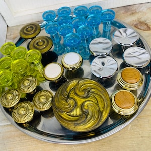 May include: A variety of decorative knobs and pulls in different styles and colors are arranged on a silver tray. The selection includes gold, silver, clear blue, and yellow-green knobs, with some featuring ornate designs and others with simple, round shapes.