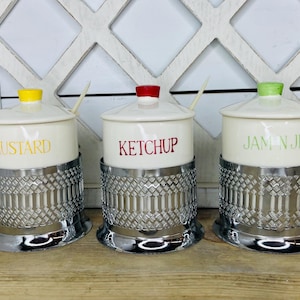 May include: Three cream-colored condiment jars with lids and spoons, labeled "MUSTARD," "KETCHUP," and "JAM N JELLY." Each jar has a different colored knob: yellow, red, and green. The jars sit in decorative silver metal holders.