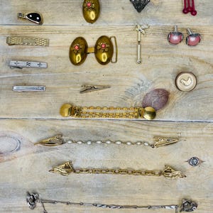 May include: A collection of vintage jewelry and accessories, including tie clips, cufflinks, a belt buckle, and a chain. The items are gold-toned, silver-toned, and feature decorative elements like pearls, rhinestones, and enamel. The pieces are arranged on a light wood surface.