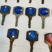 May include: Close-up view of multiple vintage keys with blue and red circular tags. Each tag displays a unique numerical code, such as 1213, 2114, 2142, 2154, 2216, 2221, and 143. The keys appear to be made of brass or a similar metal, showing signs of age and wear. The tags are firmly attached to the keys, indicating their function as room or locker keys. The numbers on the tags suggest a sequential or organizational system. The overall impression is one of antique or vintage hotel or motel keys.