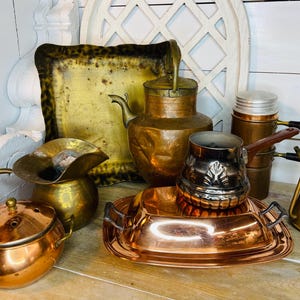 May include: An assortment of vintage copper and brass kitchenware. Includes a copper teapot, a small brass pitcher, a copper pot with a lid, a copper tray, a silver-colored coffee pot, and a brass plant mister. The items are arranged on a wooden surface.