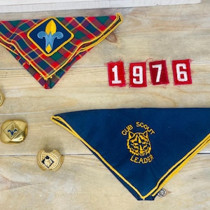 May include: Vintage Cub Scout memorabilia including two neckerchiefs, patches, and pins. One neckerchief is plaid with a fleur-de-lis patch. The other is navy blue with a tiger emblem and the words "Cub Scout Leader". The patches read "1976".