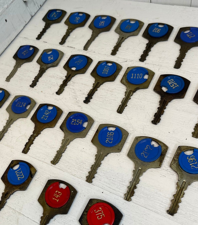 May include: A collection of vintage hotel keys with blue and red number tags. The keys are made of metal with a worn look. Numbers visible include 2221, 143, 2114, 2142, 2154, 2166, 375, 2102, 32, 2196, 742, 1007, 1110, 146, 1570, 206, and 2710. These antique keys are displayed neatly on a white surface.
