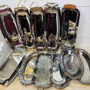 May include: A collection of vintage silver serving trays in various shapes and sizes. Some trays have handles made of wood or metal, while others have ornate, decorative edges. The trays are arranged on a light-colored wooden surface.