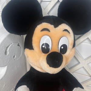May include: A plush Mickey Mouse toy with black ears, a black nose, and large white eyes. The toy is wearing red shorts with yellow buttons.