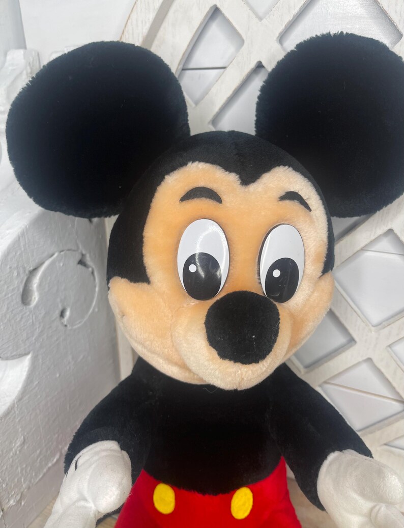 May include: A plush Mickey Mouse toy with black ears, a black nose, and large white eyes. The toy is wearing red shorts with yellow buttons.