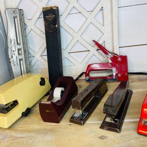 May include: A collection of vintage office supplies, including staplers, tape dispensers, and a slide rule. The staplers are in various colors, including red, yellow, and brown. The slide rule is silver. The items are arranged on a wooden surface.