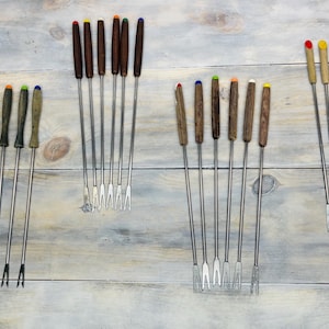 May include: A collection of fondue forks with wooden handles and stainless steel tines. The handles are various colors, including brown, green, yellow, and red. The forks are arranged on a light gray wooden surface.