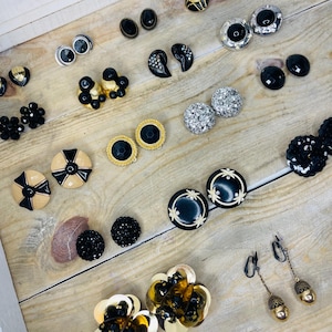May include: A collection of vintage earrings in various styles and colors, including black, gold, silver, and cream. The earrings are arranged on a light-colored wooden surface, showcasing a variety of shapes, such as studs, clip-ons, and dangling styles.