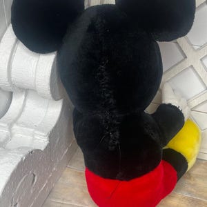 May include: A black plush Mickey Mouse toy with red shorts and yellow shoes. The toy is sitting on a wooden surface.