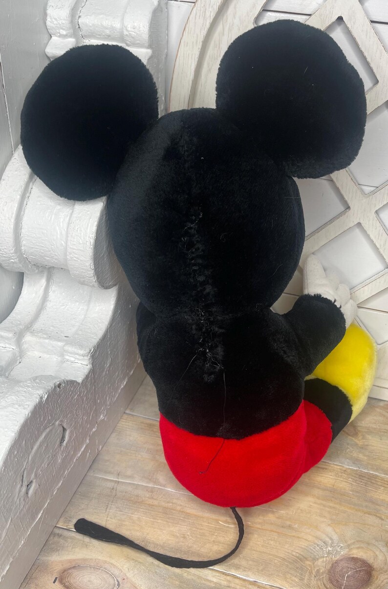 May include: A black plush Mickey Mouse toy with red shorts and yellow shoes. The toy is sitting on a wooden surface.