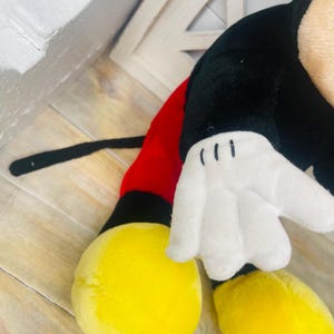 May include: A plush Mickey Mouse toy with black fur, red shorts, and yellow shoes. The toy has a white glove with three black lines on the hand.
