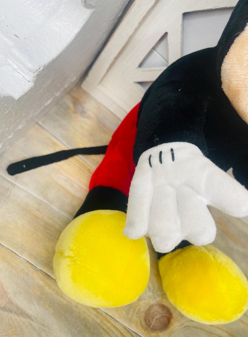 May include: A plush Mickey Mouse toy with black fur, red shorts, and yellow shoes. The toy has a white glove with three black lines on the hand.