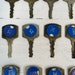 May include: Close-up view of multiple vintage hotel keys. Each key features a blue circular insert with white numbers or letters, such as 146, 156, 202, 322, 325, 742, 1001, 1007, 1213, 2114, and 2142, and N. The keys are metallic, showing signs of age and wear. The background is a white surface. These antique keys are ideal for vintage collectors, hotel enthusiasts, or those seeking unique decorative items.