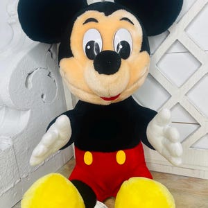 May include: A plush Mickey Mouse toy with black ears, a black nose, and yellow shoes. The toy is wearing red shorts with two yellow buttons.