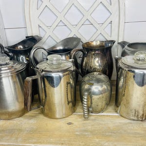 May include: Collection of vintage metal coffee pots, pitchers, and a small sugar bowl. The coffee pots have glass knob lids and wooden handles. The pitchers are silver and dark metal. The items are arranged on a wooden surface.