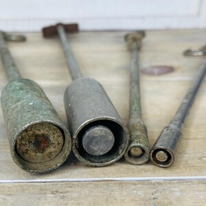 Peut inclure: Quatre outils anciens en m&eacute;tal avec de longs manches et des extr&eacute;mit&eacute;s cylindriques ou coniques. Les outils ont un aspect vieilli et patin&eacute;, avec un m&eacute;lange de patines argent&eacute;es et verd&acirc;tres. Les outils sont dispos&eacute;s sur une surface en bois clair.