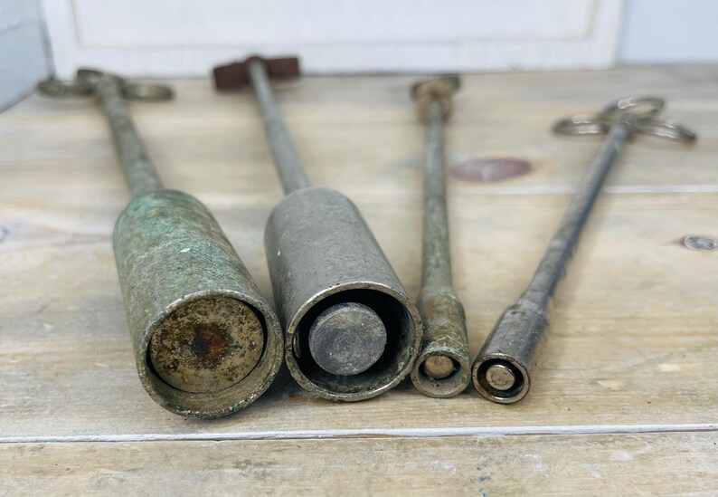 Peut inclure: Quatre outils anciens en m&eacute;tal avec de longs manches et des extr&eacute;mit&eacute;s cylindriques ou coniques. Les outils ont un aspect vieilli et patin&eacute;, avec un m&eacute;lange de patines argent&eacute;es et verd&acirc;tres. Les outils sont dispos&eacute;s sur une surface en bois clair.